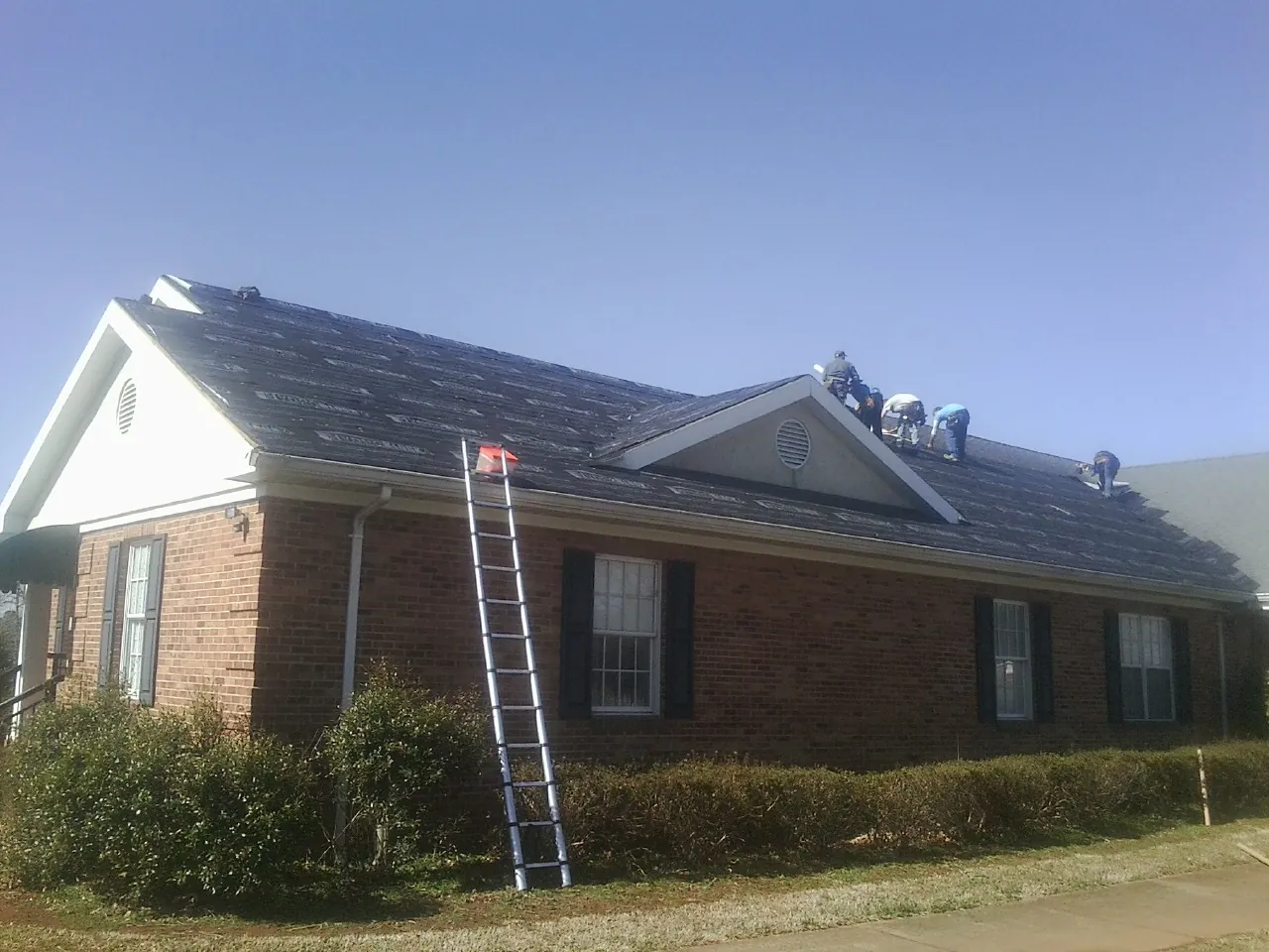 Crew tearing shingles off a brick ranch roof with a ladder set — Emergency Roof Repair work by our crew in East Troy