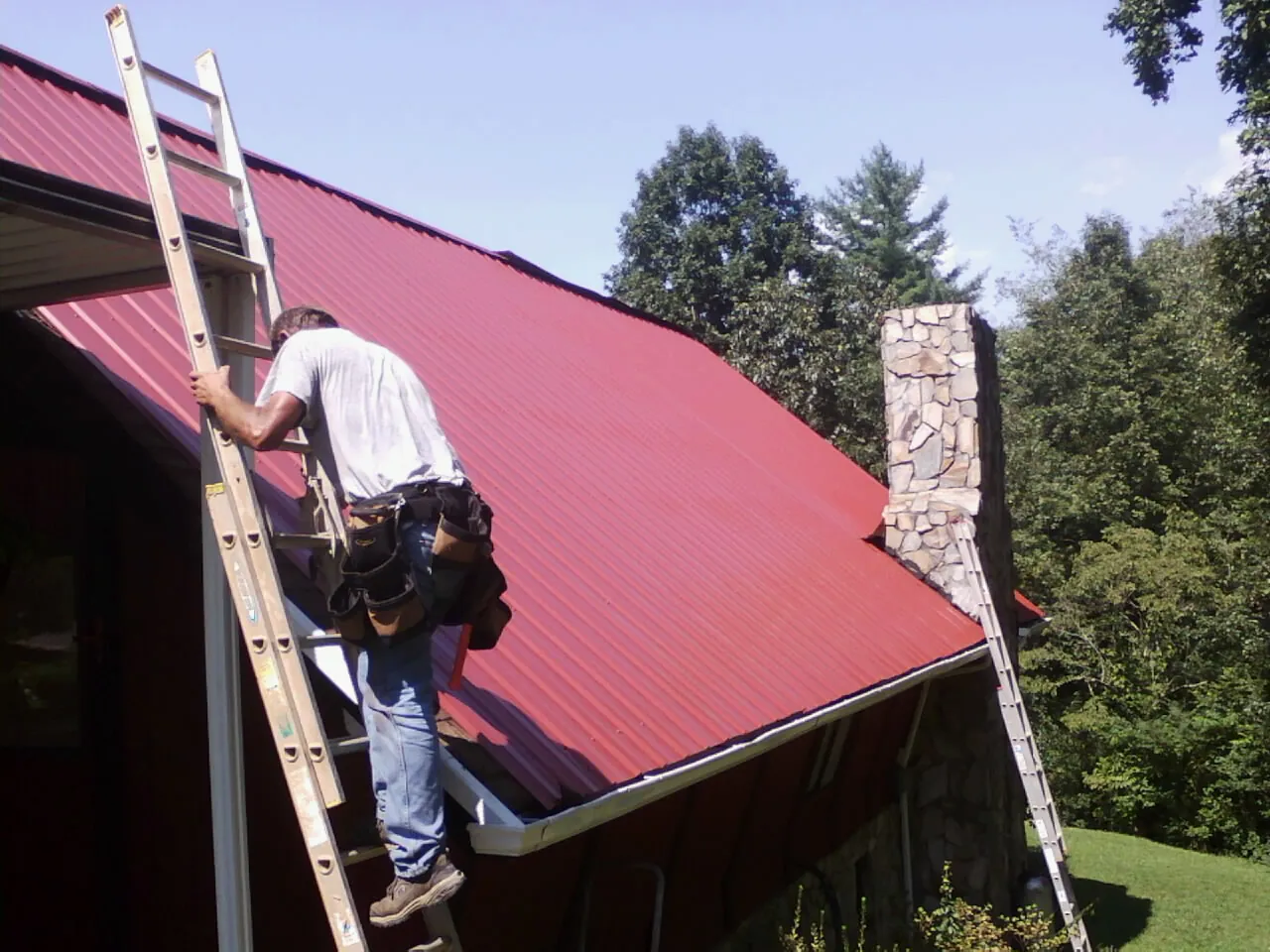 Roofer on a ladder beside a red metal roof and stone chimney — Soffit & Fascia Repair by Roofing Brothers in East Troy